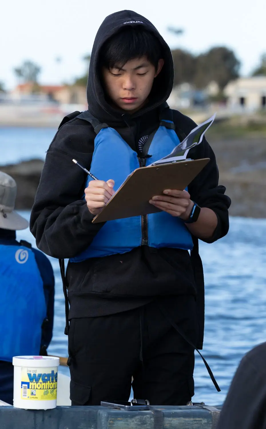 Chadwick Upper School student in a hoodie writes notes on a clipboard while on a kayak in the ocean.