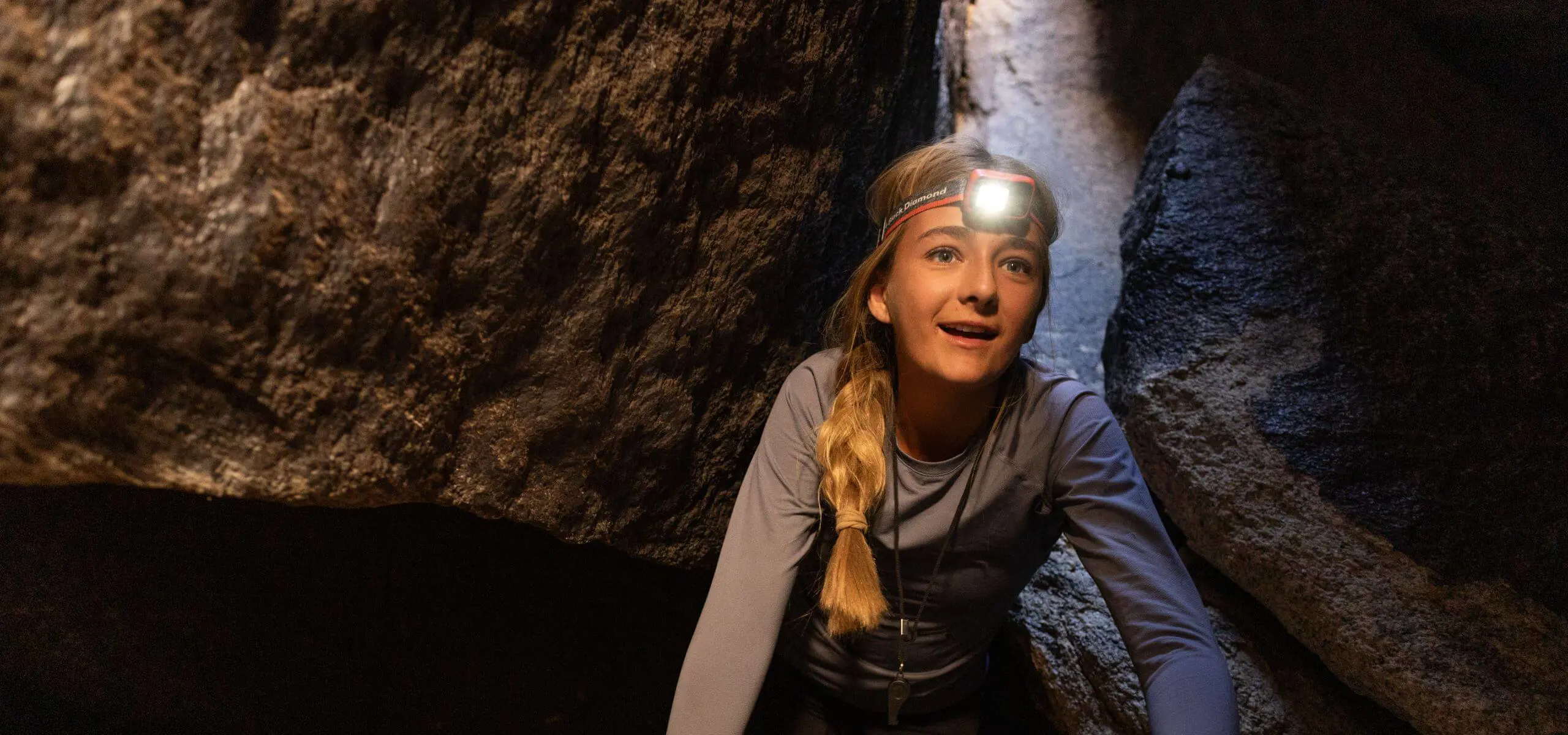 Middle School student wearing a lit headlamp smiles and looks up while scrambling in a rock cave in Joshua Tree National Park.