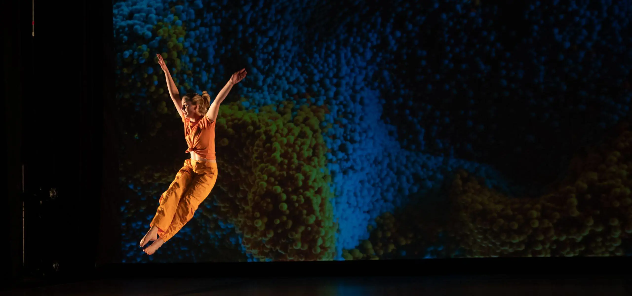 Chadwick dancer takes a leap wearing orange pants and top in front of a blue background while in stage.