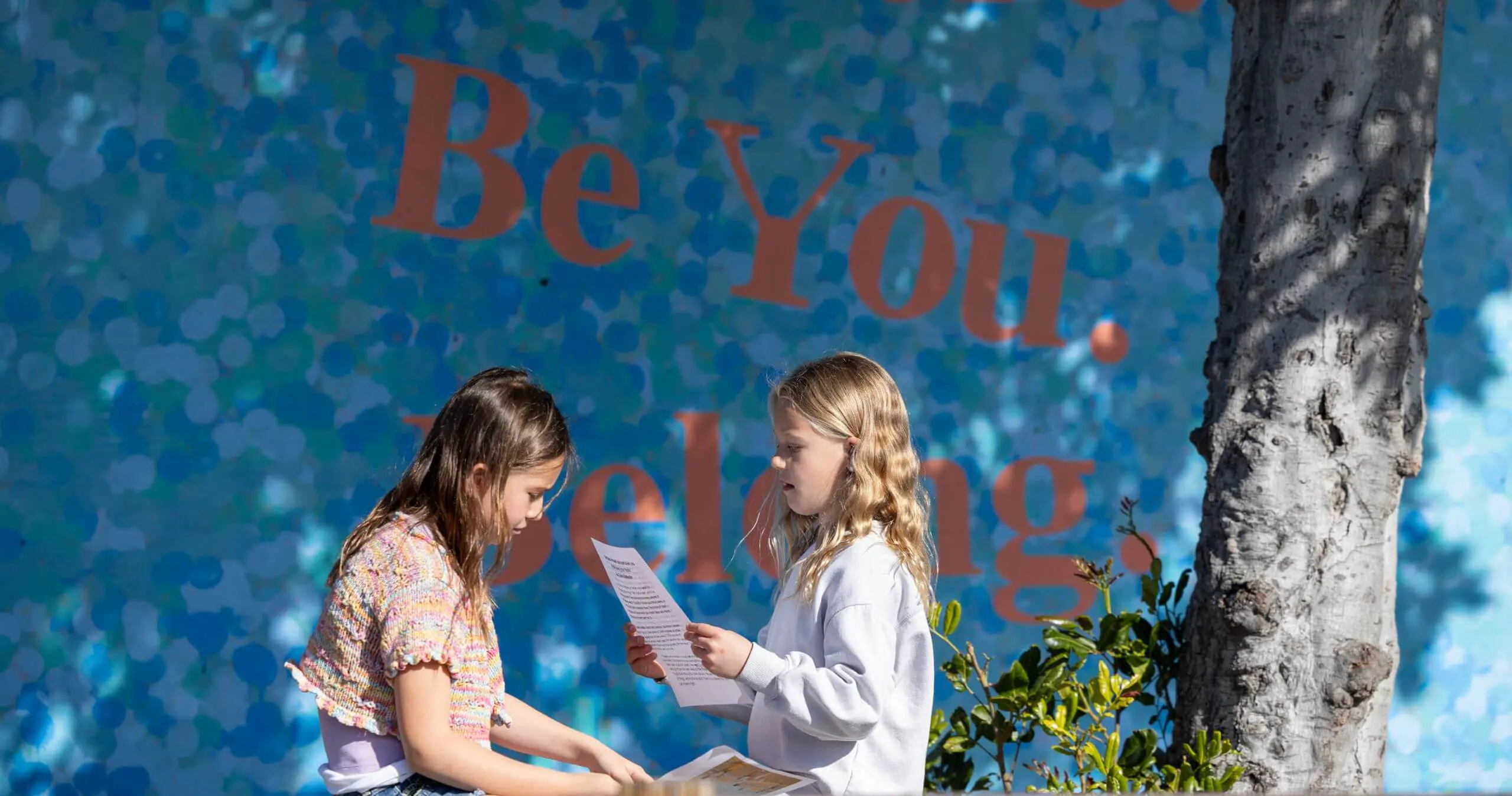 Two students collaborate in front of a mural that reads, “Be Here. Be you. Belong”